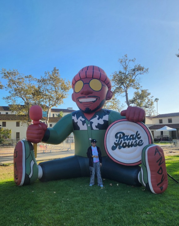 Kk is wearing a cap with a black sweater and is standing in front of a 15ft+ blow up character of Anderson Paak