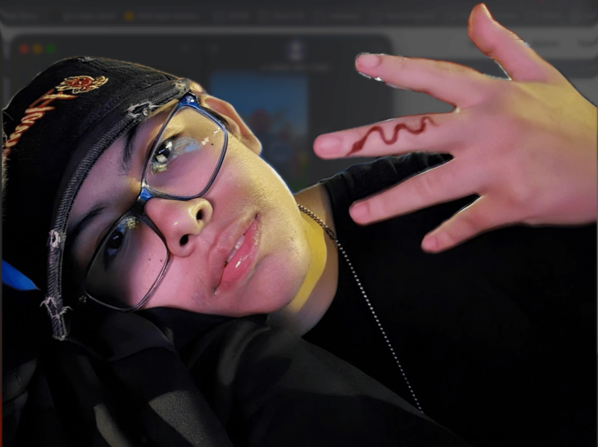 Kk is wearing a cap and black rimmed glasses with her hand up and a red snake design is on her finger